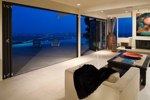 Open corner doors connecting a living room to a pool terrace at dusk.